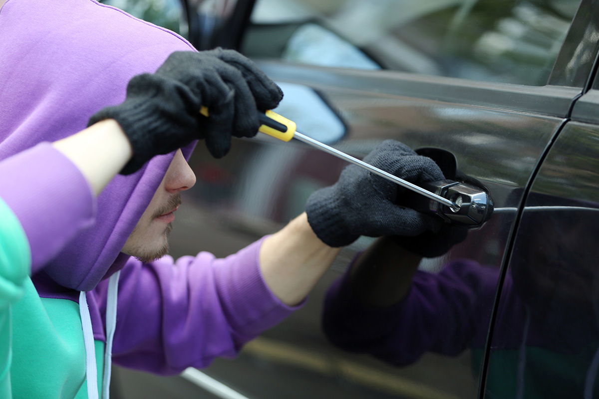 A man in a purple and turquois hoody breaking into a black car with a screwdriver.