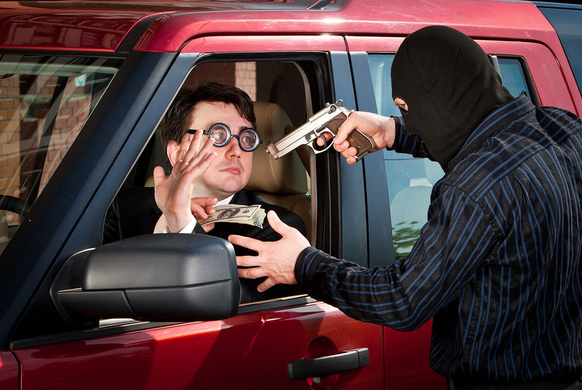 A man wearing a business suit and glasses is being robbed at gun point by a man in a black mask while he sits in a red SUV. 